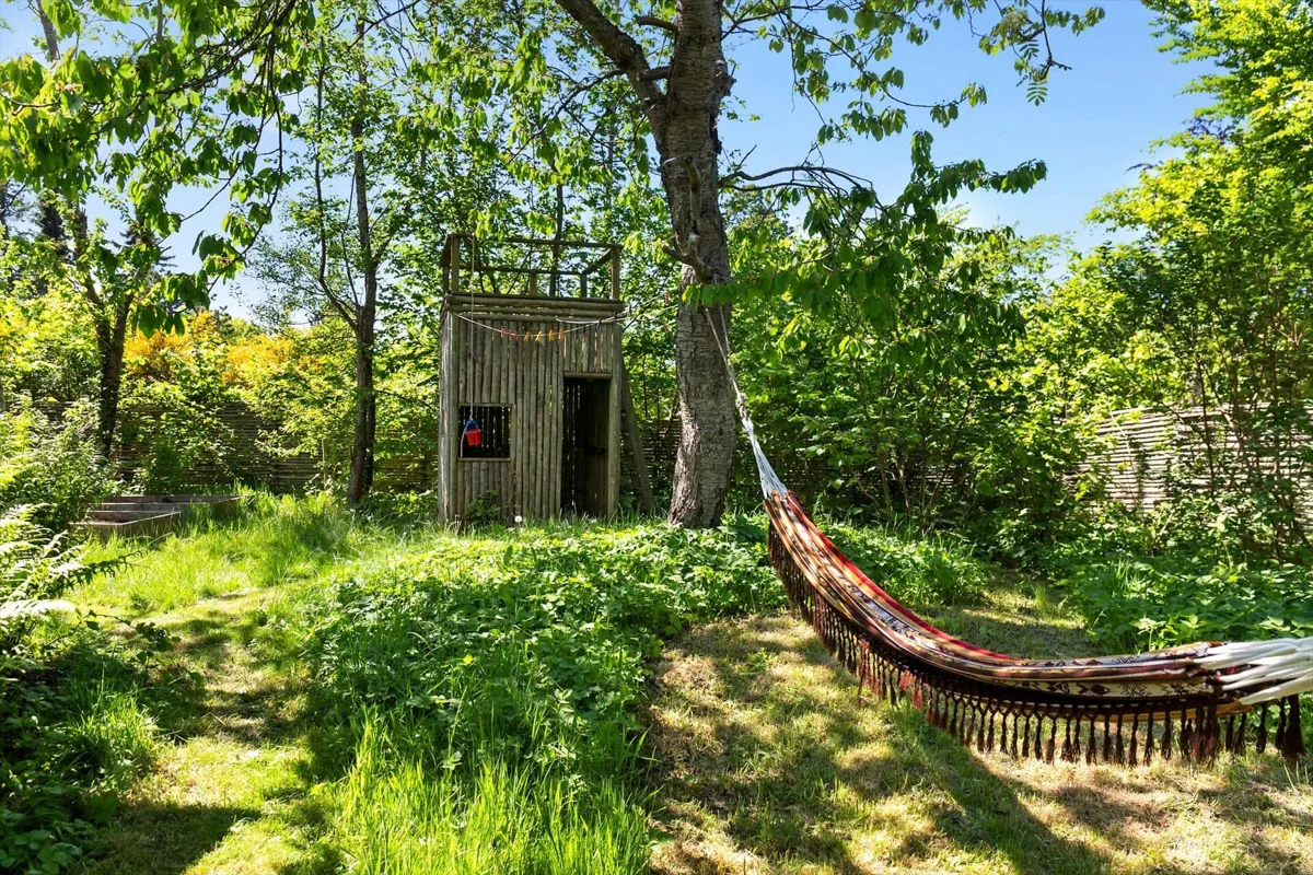 Grönskande trädgård i solsken med en lekstuga i trä och en randig hängmatta uppspänd i ett träd